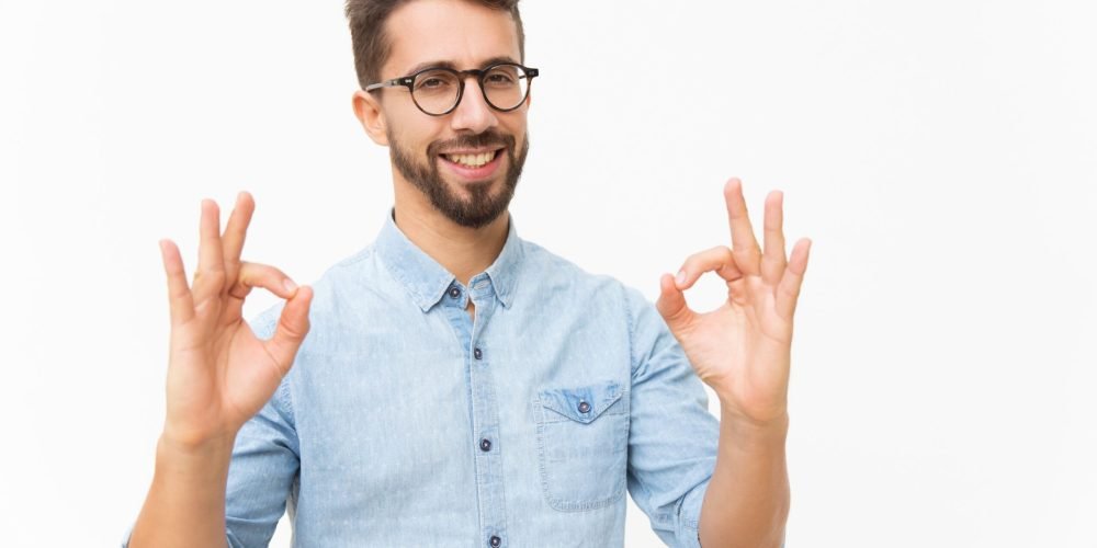 Happy joyful guy showing OK gesture Happy joyful guy showing OK gesture with both hands. Handsome young man in casual shirt and glasses standing isolated over white background. Customer satisfaction concept