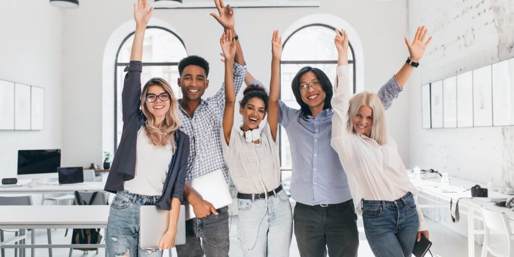 International tired students celebrating end of exams and laughing in lecture hall. Happy freelance programmers done with long project and posing with smile, holding laptops.